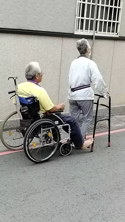 彰化市這對長者相互扶持走在街頭，前者拄著四腳枴杖，用一條繩子拉著坐輪椅的長者，緩...