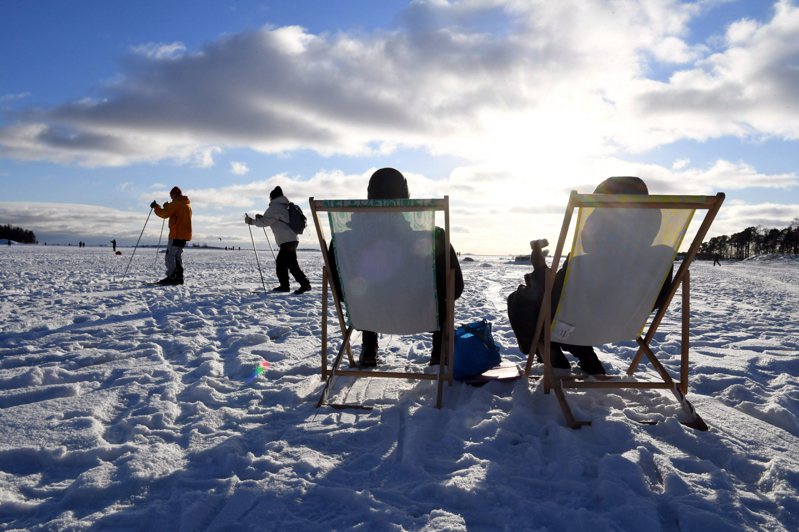 芬蘭名列全球最幸福國家第一，圖為人們坐在赫爾辛基海濱冰上，其他人滑雪經過。(美聯社)