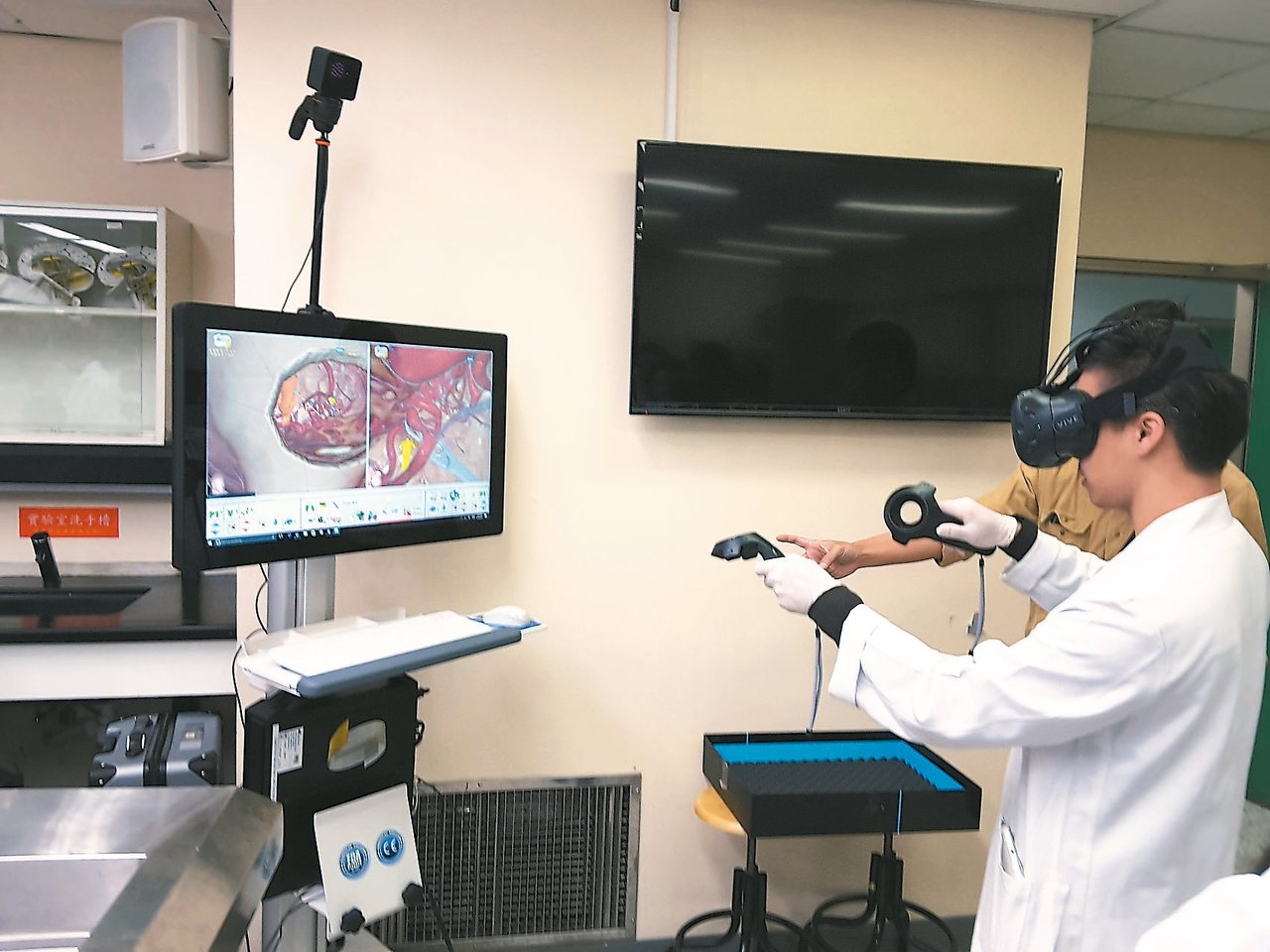 醫學生正使用頭戴顯示器與數據搖桿感受實境動脈瘤虛擬手術，在VR鏡頭裡看到的就和電腦螢幕上一樣，如同置身於手術場景，跟執刀醫師看到的一樣, 可以從多角度及多模組嘗試手術過程直到滿意為止，因為腦部精細手術如同太空飛行模擬，只許成功不容些許差池。<br />圖／湯其暾醫師提供
