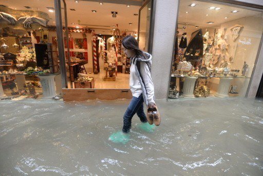 一名女子在威尼斯市區拎著鞋涉水前行，旁邊的服飾店門口架起閘門擋水。 （美聯社）