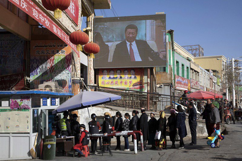 中國對新疆維吾爾穆斯林進行高壓灌輸計畫，圖中民眾進和田市集前需先通過安檢站，上方螢幕為中國國家主席習近平。美聯社