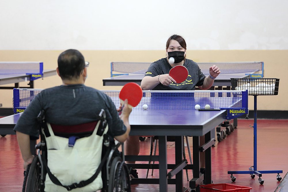 為了與身障者維持同樣高度，蔡貴蘭教練坐在椅子上陪伴肢體障礙者桌球推廣營的學員練球...