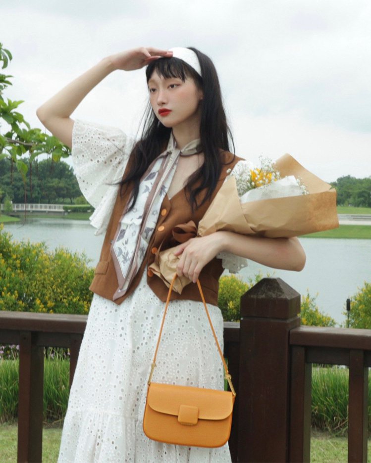時裝模特兒Kiki示範背上橘色的Koa方釦包，搭配白色鏤空洋裝、棕色背心和印花絲...