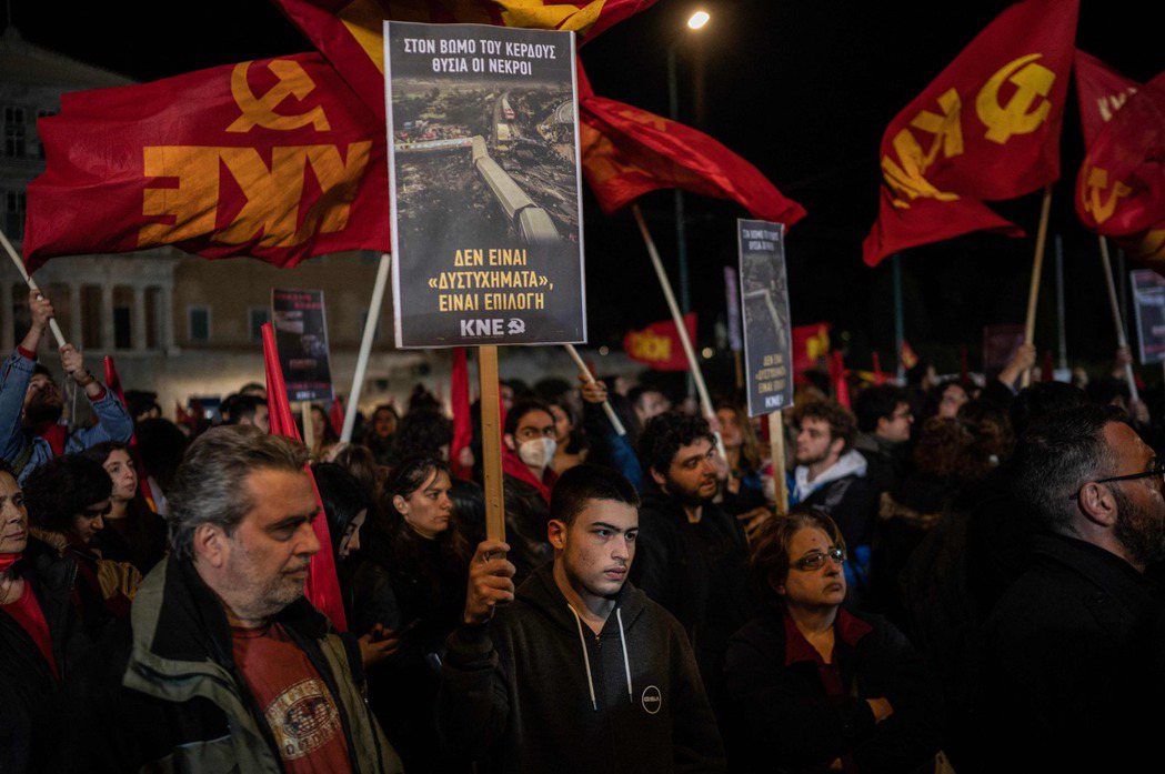 火車對撞事故引發希臘全國抗議，雅典的一名抗爭者在希臘國會前舉著一張海報，上面寫著...