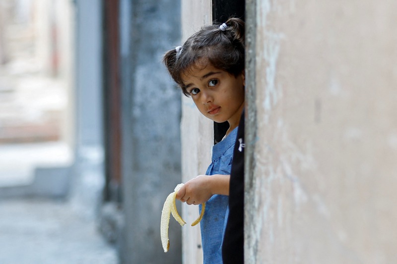 一名巴勒斯坦女童從加薩南部的難民收容所探頭往外望，手裡還拿著香蕉。路透