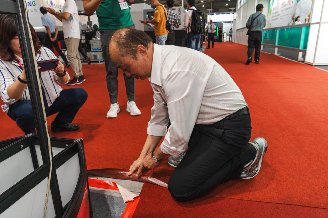 輝祐董事長王崑池在展館中蹲下並掀起地板上的地毯示範，強調為了通過ISO 2012...