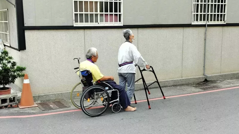 彰化市這對長者相互扶持走在街頭，前者拄著四腳枴杖，用一條繩子拉著坐輪椅的長者，緩...